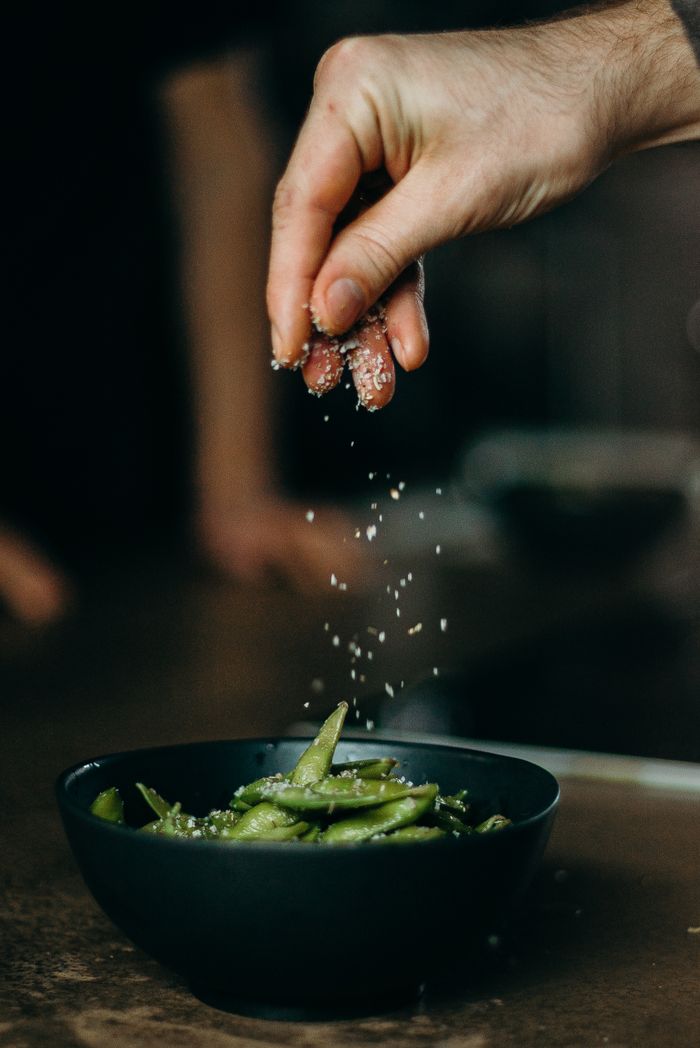 A hand sprinkles salt over a bowl of fresh edamame beans, showcasing a close-up culinary moment.